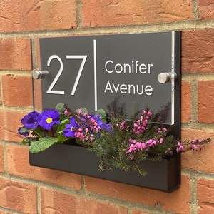 May include: A black acrylic house number sign with white lettering. The sign reads "27 Conifer Avenue". The sign is mounted on a brick wall with a black planter box underneath.