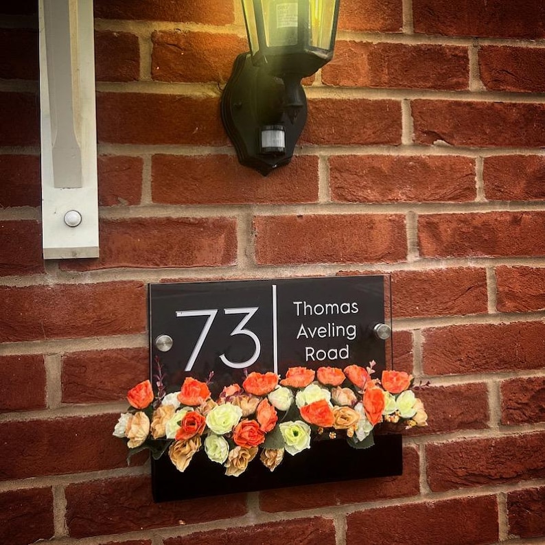 May include: Black acrylic house number sign with white numbers and text. The sign reads "73 Thomas Aveling Road". The sign is mounted on a brick wall with a light fixture above it. A row of artificial flowers is in front of the sign.