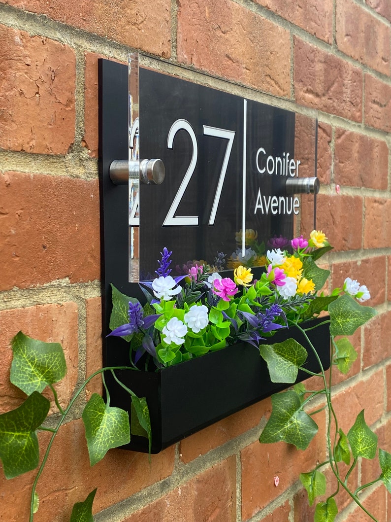May include: Black acrylic house number sign with white lettering, displaying the number 27 and the street name Conifer Avenue. The sign is mounted on a brick wall with a black planter box underneath, filled with artificial flowers.