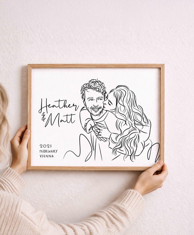 May include: A framed line art illustration featuring a couple with the text Heather & Matt, 2023, February, and Vienna. The artwork is rendered in black ink on a white background, enclosed in a light-coloured wooden frame with a simple border.