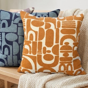 May include: Three decorative throw pillows with geometric patterns in shades of purple, blue, and orange. The pillows are arranged on a wooden bench, offering a stylish accent for home decor. The pillows have a textured appearance.