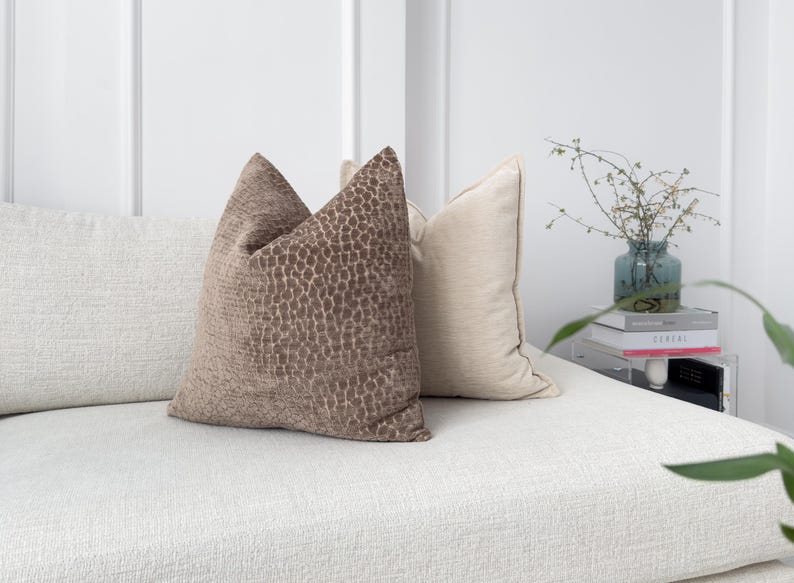 Puede incluir: Dos cojines decorativos sobre un sof&aacute; blanco texturizado. Un coj&iacute;n es de color beige liso, y el otro es de color marr&oacute;n con un patr&oacute;n texturizado. Los cojines est&aacute;n colocados contra una pared blanca.
