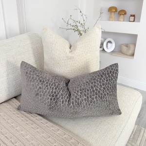 Puede incluir: Un coj&iacute;n decorativo gris con un patr&oacute;n texturizado descansa sobre un sof&aacute; color crema, junto a un coj&iacute;n color crema con una textura similar. Una manta de punto beige est&aacute; drapeada sobre el sof&aacute;. Los cojines a&ntilde;aden un toque de elegancia a la sala de estar.