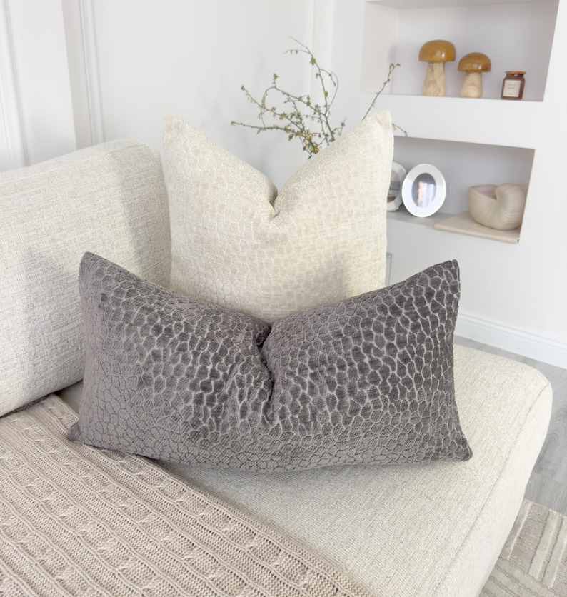 Puede incluir: Un coj&iacute;n decorativo gris con un patr&oacute;n texturizado descansa sobre un sof&aacute; color crema, junto a un coj&iacute;n color crema con una textura similar. Una manta de punto beige est&aacute; drapeada sobre el sof&aacute;. Los cojines a&ntilde;aden un toque de elegancia a la sala de estar.