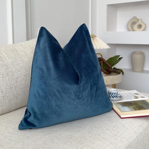 May include: A teal blue velvet pillow on a white couch. The pillow is square and has a smooth, soft texture.