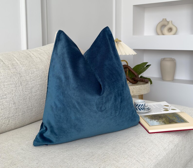 May include: A teal blue velvet pillow on a white couch. The pillow is square and has a smooth, soft texture.