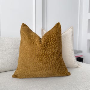 K&ouml;nnte beinhalten: Ein senfgelbes Kissen mit einem strukturierten, geometrischen Muster. Das Kissen liegt auf einem wei&szlig;en Sofa.
