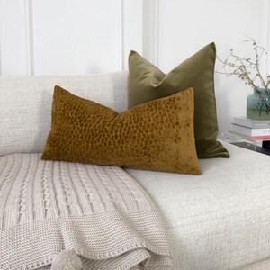 K&ouml;nnte beinhalten: Ein wei&szlig;es Sofa mit einer beigefarbigen Strickdecke und zwei Kissen. Ein Kissen ist aus olivgr&uuml;nem Samt und das andere aus braunem Samt mit einem strukturierten Muster.