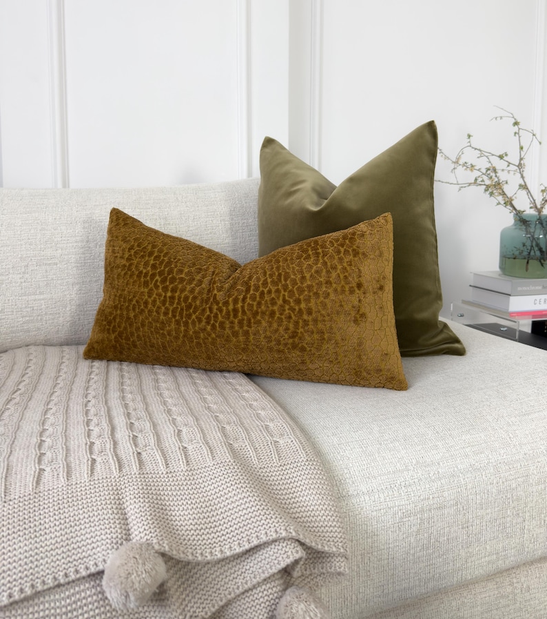 K&ouml;nnte beinhalten: Ein wei&szlig;es Sofa mit einer beigefarbigen Strickdecke und zwei Kissen. Ein Kissen ist aus olivgr&uuml;nem Samt und das andere aus braunem Samt mit einem strukturierten Muster.