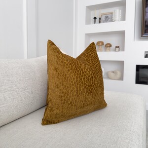 K&ouml;nnte beinhalten: Ein braunes Samtkissen mit einem strukturierten Muster. Das Kissen liegt auf einem wei&szlig;en Sofa.