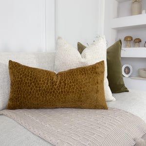 Puede incluir: Una disposici&oacute;n decorativa de cojines en varios colores y texturas. El coj&iacute;n principal es rectangular, marr&oacute;n y texturizado. Otros cojines son blancos y verde oliva, creando un ambiente acogedor y elegante.