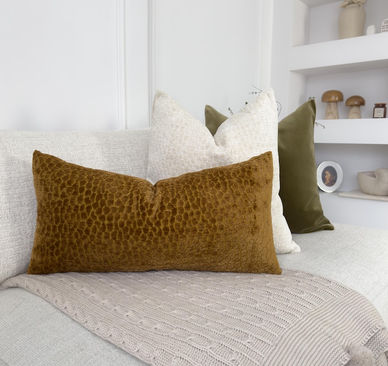 Puede incluir: Una disposici&oacute;n decorativa de cojines en varios colores y texturas. El coj&iacute;n principal es rectangular, marr&oacute;n y texturizado. Otros cojines son blancos y verde oliva, creando un ambiente acogedor y elegante.