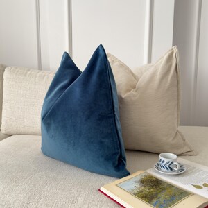 May include: A blue velvet pillow on a white couch with a beige pillow behind it. A book and a teacup are on the couch.