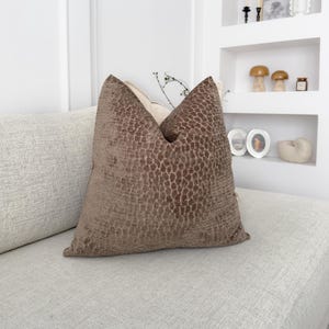 Puede incluir: Un coj&iacute;n decorativo marr&oacute;n con un patr&oacute;n texturizado, colocado sobre un sof&aacute; de color claro. El coj&iacute;n tiene un aspecto suave y afelpado y a&ntilde;ade un toque de elegancia al espacio habitable. El dise&ntilde;o del coj&iacute;n recuerda a la piel de un reptil.