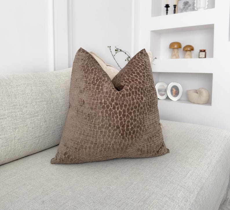 Puede incluir: Un coj&iacute;n decorativo marr&oacute;n con un patr&oacute;n texturizado, colocado sobre un sof&aacute; de color claro. El coj&iacute;n tiene un aspecto suave y afelpado y a&ntilde;ade un toque de elegancia al espacio habitable. El dise&ntilde;o del coj&iacute;n recuerda a la piel de un reptil.