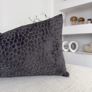Op de afbeelding: Een donkergrijs decoratief kussen met een textuur, honingraatachtig patroon. Het kussen ligt op een lichtgekleurd oppervlak, met een witte plank en decoratieve items op de achtergrond.