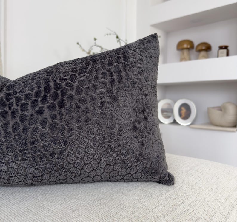 Op de afbeelding: Een donkergrijs decoratief kussen met een textuur, honingraatachtig patroon. Het kussen ligt op een lichtgekleurd oppervlak, met een witte plank en decoratieve items op de achtergrond.