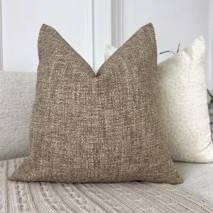 May include: A brown throw pillow with a textured fabric. The pillow is sitting on a cream-coloured knitted throw blanket.