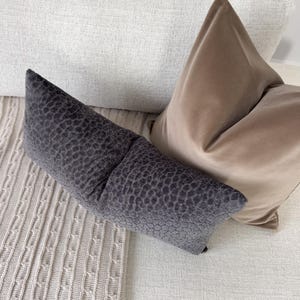 Op de afbeelding: Een close-up van drie decoratieve kussens in neutrale tinten. Een lang, donkergrijs kussen met een textuurpatroon staat op de voorgrond. Een tan fluwelen kussen en een wit kussen staan erachter. De kussens liggen op een lichtgekleurde bank.