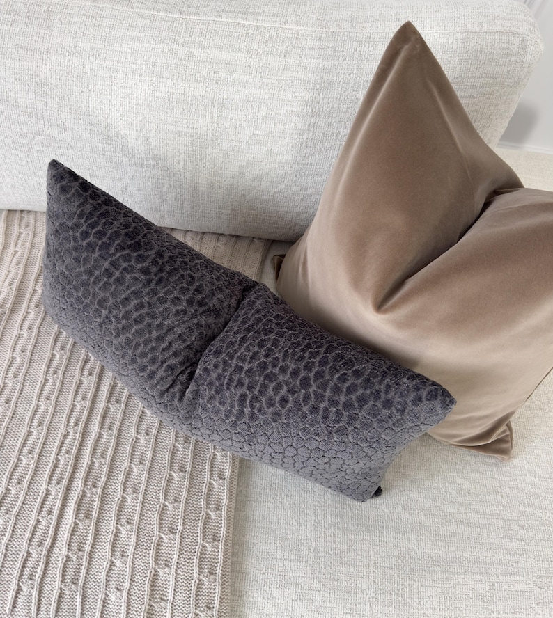 Op de afbeelding: Een close-up van drie decoratieve kussens in neutrale tinten. Een lang, donkergrijs kussen met een textuurpatroon staat op de voorgrond. Een tan fluwelen kussen en een wit kussen staan erachter. De kussens liggen op een lichtgekleurde bank.