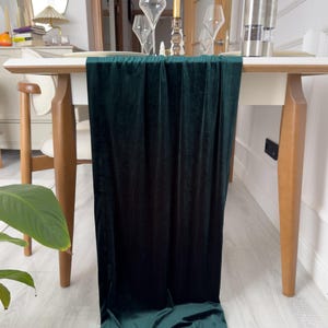 May include: A deep emerald green velvet table runner cascading over a white table. The runner is made of a soft, luxurious fabric, adding elegance to a dining setting. The table is set with wine glasses and a candle.