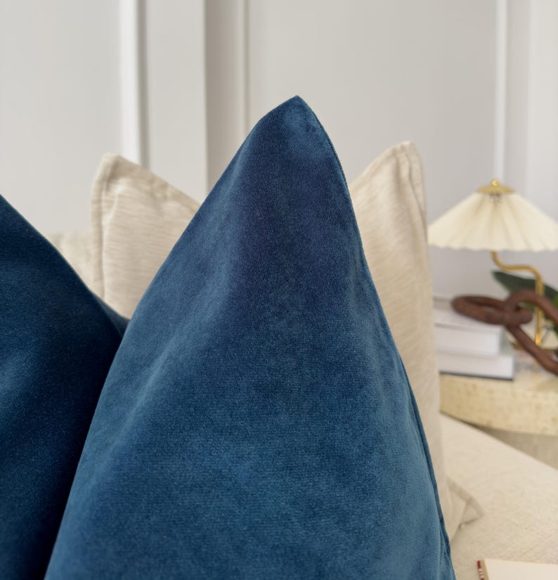 May include: Close-up of a teal blue velvet pillow with a soft, plush texture. The pillow is positioned on top of a cream-colored pillow.
