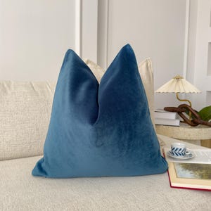May include: A teal blue velvet pillow on a cream colored couch. The pillow is square shaped and has a simple design.