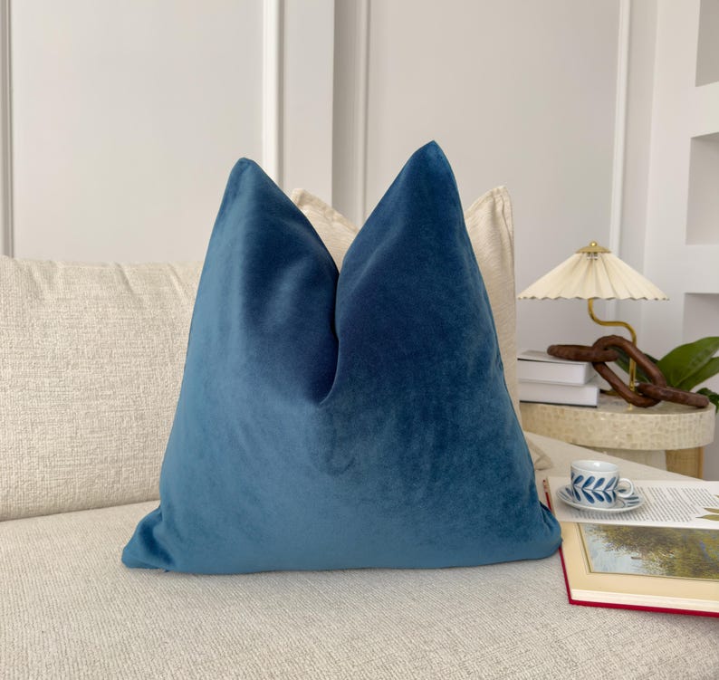 May include: A teal blue velvet pillow on a cream colored couch. The pillow is square shaped and has a simple design.