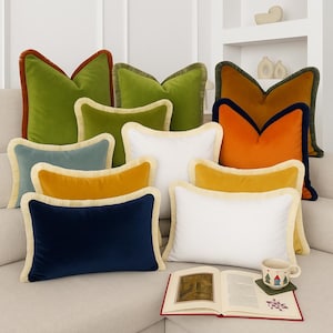 May include: A collection of decorative throw pillows in various colors, including green, orange, blue, yellow, and white. Each pillow has a contrasting border and fringe trim. A book and a mug with a house design are also visible.