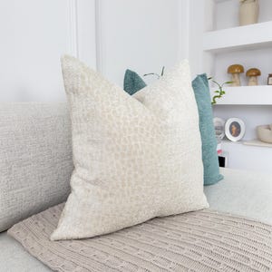 Puede incluir: Una colecci&oacute;n de cojines decorativos en tonos neutros. El coj&iacute;n delantero es de color beige claro con un patr&oacute;n texturizado. Detr&aacute;s hay un coj&iacute;n turquesa. Una manta de punto de color marr&oacute;n claro est&aacute; en primer plano.