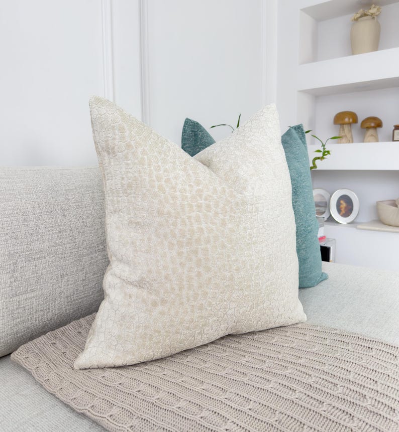 Puede incluir: Una colecci&oacute;n de cojines decorativos en tonos neutros. El coj&iacute;n delantero es de color beige claro con un patr&oacute;n texturizado. Detr&aacute;s hay un coj&iacute;n turquesa. Una manta de punto de color marr&oacute;n claro est&aacute; en primer plano.