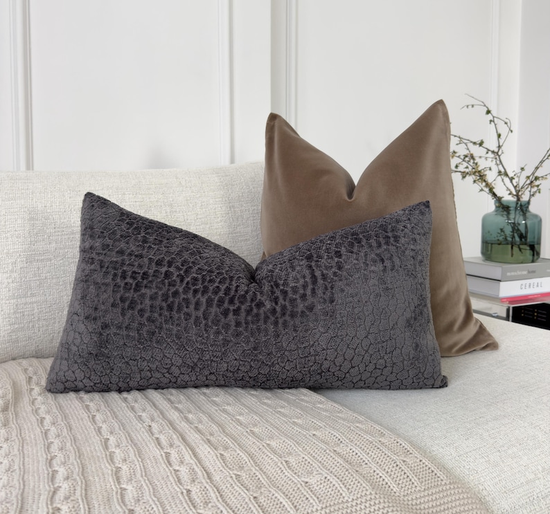 Puede incluir: Dos cojines decorativos sobre un sof&aacute; blanco. El coj&iacute;n delantero es de color gris oscuro con un patr&oacute;n texturizado. El coj&iacute;n trasero es de color beige liso. Una manta color crema est&aacute; sobre el sof&aacute;.