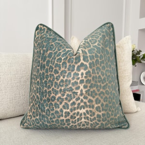 Puede incluir: Un cojín decorativo con un estampado de leopardo en color verde azulado y dorado. El cojín tiene un borde verde azulado y está colocado sobre un sofá de color neutro. El cojín tiene forma cuadrada y añade un toque de lujo a cualquier espacio habitable.