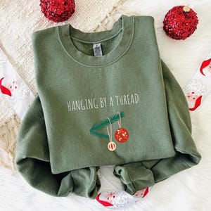 May include: An olive green sweatshirt with the phrase "HANGING BY A THREAD" printed on the front. The design includes a sprig of pine needles and two Christmas ornaments. Red sequined Christmas ornaments and Santa Claus ribbon are in the background.