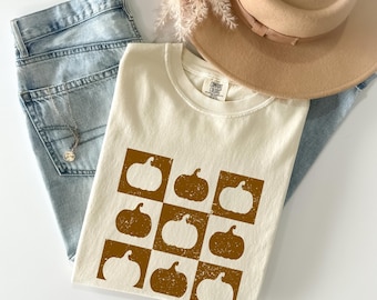 May include: Cream-colored t-shirt with a brown pumpkin pattern in a checkerboard design. The shirt is paired with light blue jeans, a tan hat, and a necklace. The shirt has a Comfort Colors tag.