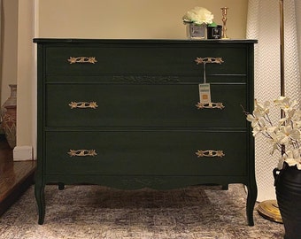 May include: A dark green dresser with three drawers and gold-toned hardware. The dresser has a carved design on the front and sits on a patterned rug.