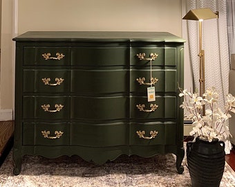 May include: A dark green dresser with five drawers and gold handles. The dresser has a curved front and sits on a patterned rug. A black vase with white flowers sits on the rug next to the dresser.