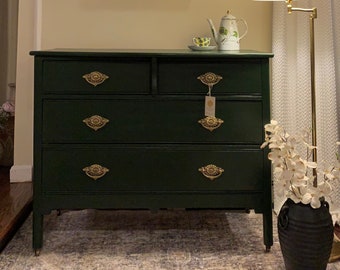 May include: A dark green dresser with four drawers and ornate gold hardware. Above the dresser hangs a framed painting of fruit. A lamp with a pleated shade and a vase of white flowers are also in the room.