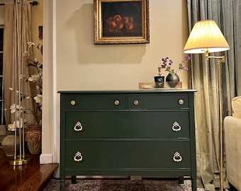 May include: A dark green dresser with three drawers and gold hardware. The dresser has a traditional style and is sitting on a patterned rug in front of a gold floor lamp.