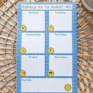 May include: A blue and white weekly planner with the text "BAHALA KA SA BUHAY MO" at the top. Each day of the week is listed with a blank space for notes, and decorated with yellow sun illustrations. The planner is on a woven mat.