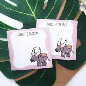 May include: Two square sticky note pads with a pink border and a white center. Each pad features a cartoon illustration of a grey carabao wearing a flower crown and a tan saddle, with the text THINGS TO CARABAO printed at the top of the page.