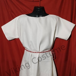 Può includere: Una tunica bianca a maniche corte con una cintura in corda rossa e bianca. La tunica è su un manichino su uno sfondo rosso. Il testo "Viking Costume" è visibile nell'angolo in basso a sinistra dell'immagine.
