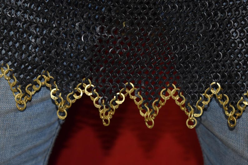 May include: Close-up of a black chainmail garment with a decorative gold-colored trim along the bottom edge. The chainmail is made of small, interlocking metal rings. The trim is made of gold-colored rings.