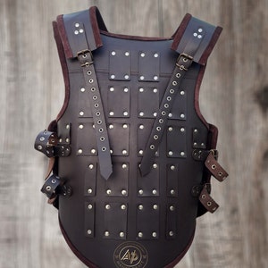 May include: A brown leather armour vest with metal rivets and straps. The vest has a rounded bottom and a logo that says "A.P. Handicraft".