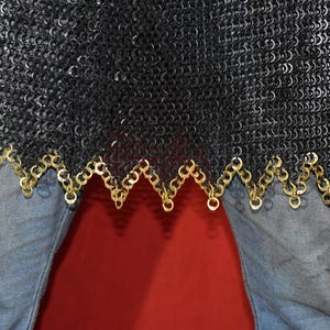May include: Close-up of a black chainmail garment with a decorative gold-colored trim. The chainmail is made of small, interlocking metal rings. The garment is worn over blue jeans.