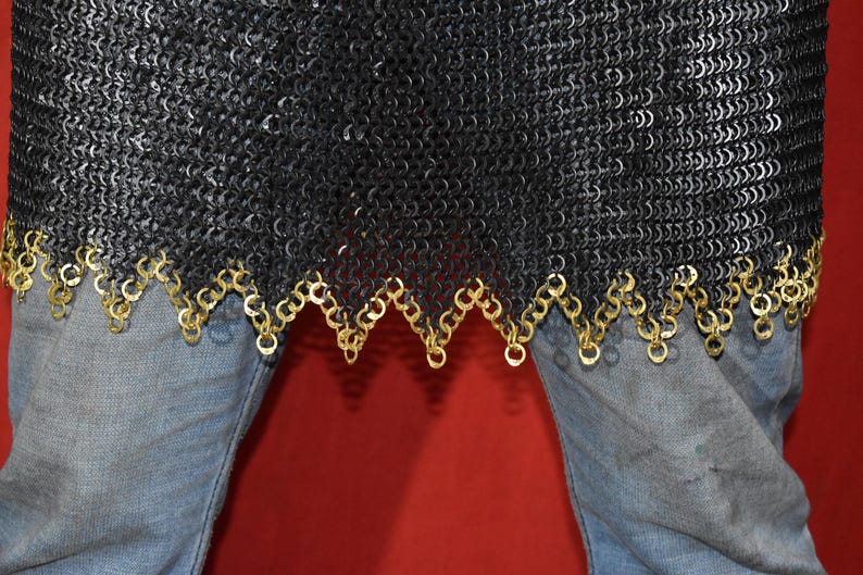 May include: Close-up of a black chainmail garment with a decorative gold-colored trim. The chainmail is made of small, interlocking metal rings. The garment is worn over blue jeans.