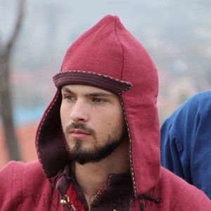 May include: A red wool hat with a pointed crown and ear flaps. The hat has a brown trim around the brim and a white stitching detail. The hat is worn by a person with a beard.