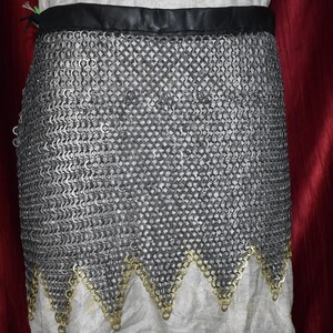 May include: A chainmail skirt with a black leather waistband and a decorative gold-colored trim along the bottom edge. The skirt is made of small, interlocking metal rings, creating a flexible, protective garment. The skirt is shown against a white fabric background.