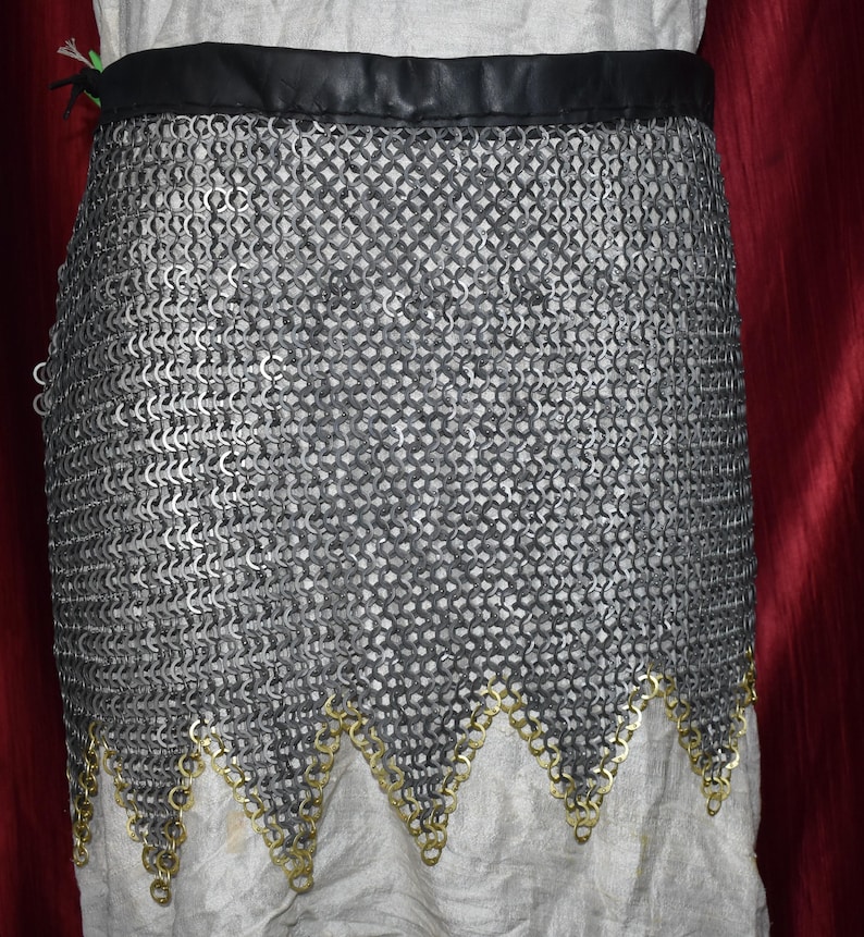 May include: A chainmail skirt with a black leather waistband and a decorative gold-colored trim along the bottom edge. The skirt is made of small, interlocking metal rings, creating a flexible, protective garment. The skirt is shown against a white fabric background.
