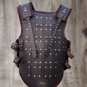 May include: A brown leather armour vest with metal rivets and straps. The vest has a rounded bottom and a logo that says "A.P. Handicraft".